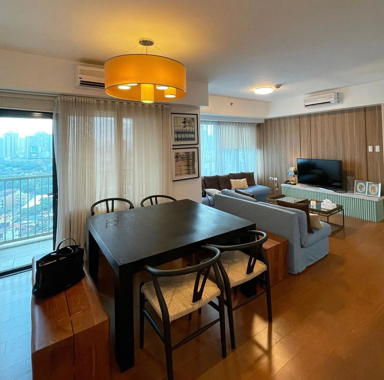 Modern living room with a dining table, sofas, large windows, and a wall-mounted TV.